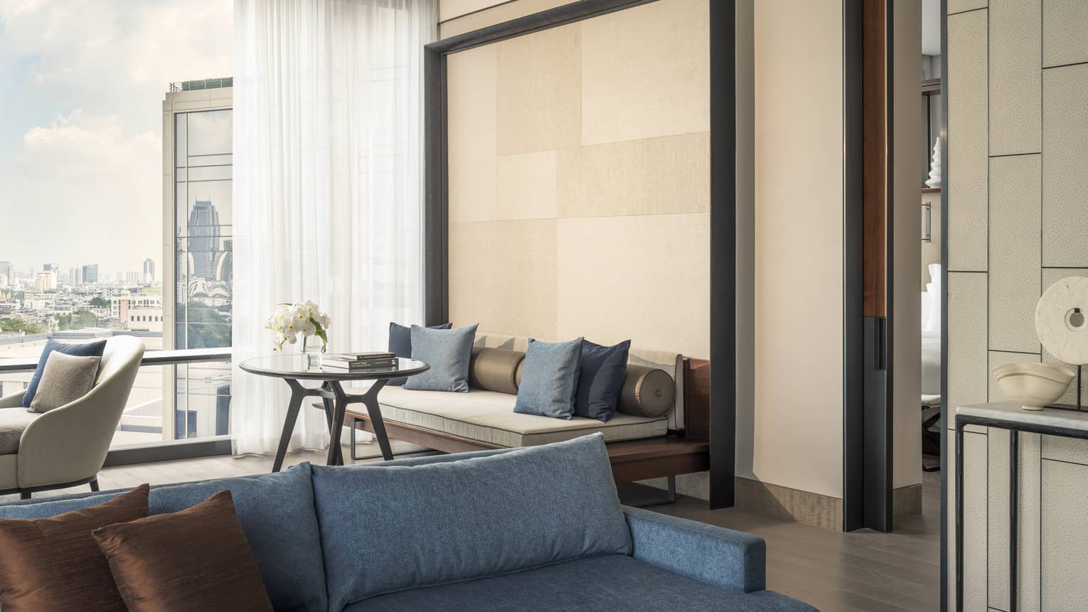 Hotel suite living area with blue sofa in forefront, sofa against tall beige accent wall, city view