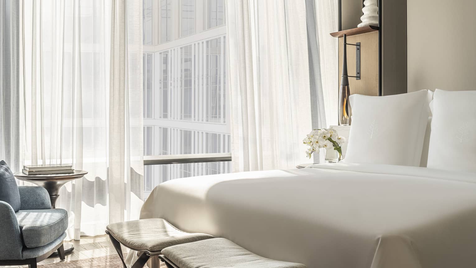 Hotel room with white bed and two benches at foot, facing blue chair, off-white wall by curtained window