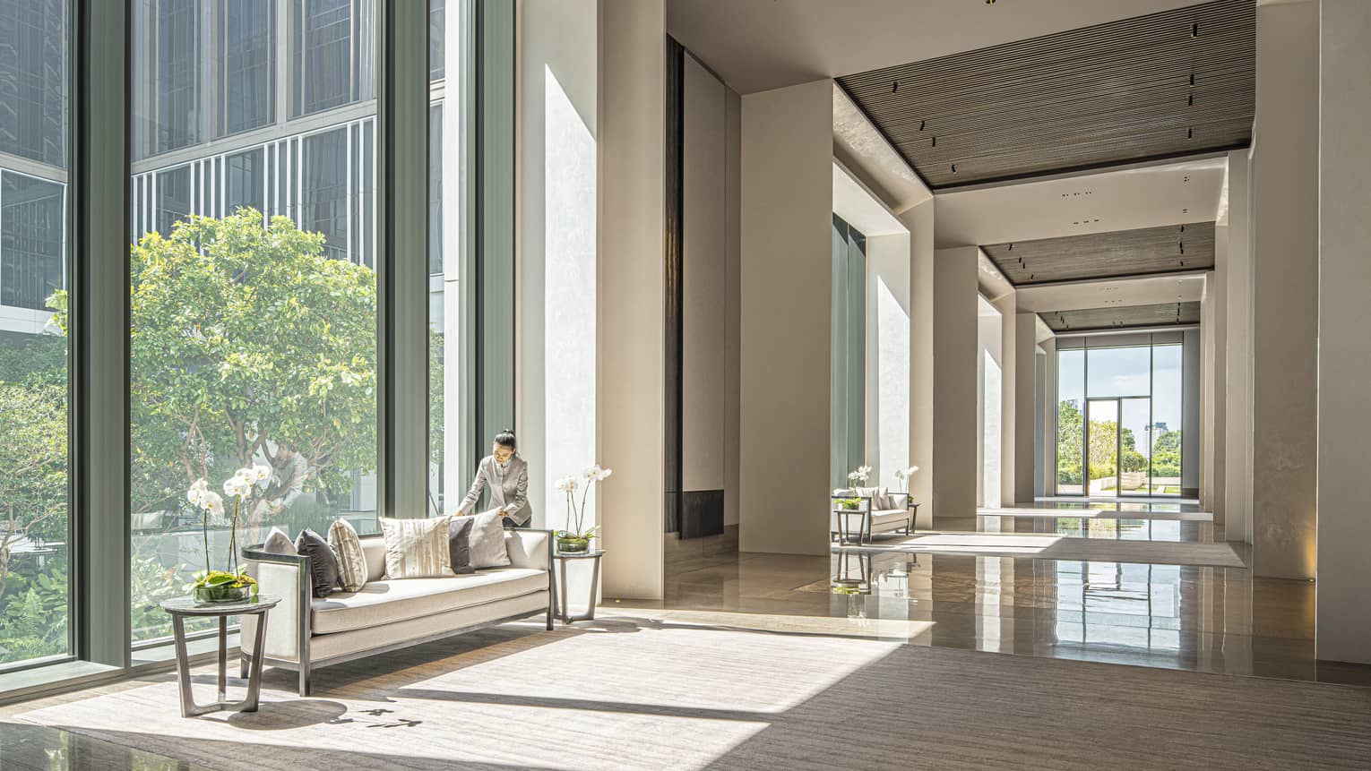 Staff member fluffs the couch pillows in the Ballroom Foyer with cream-coloured carpeting and furniture, floor-to-ceiling windows