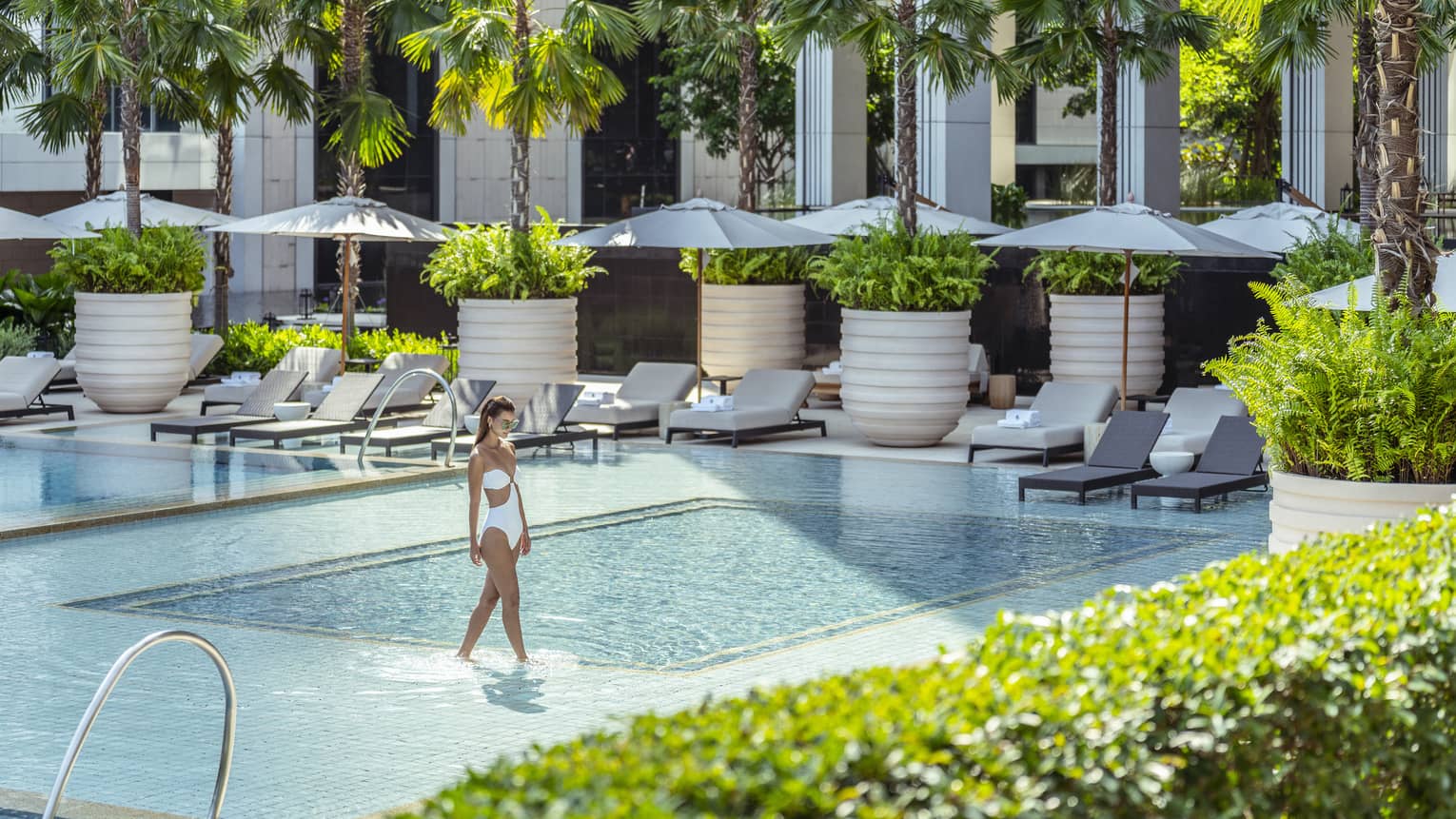 Woman in white bathing suit wades in outdoor pool surrounded by lounge chairs with umbrellas and palm trees