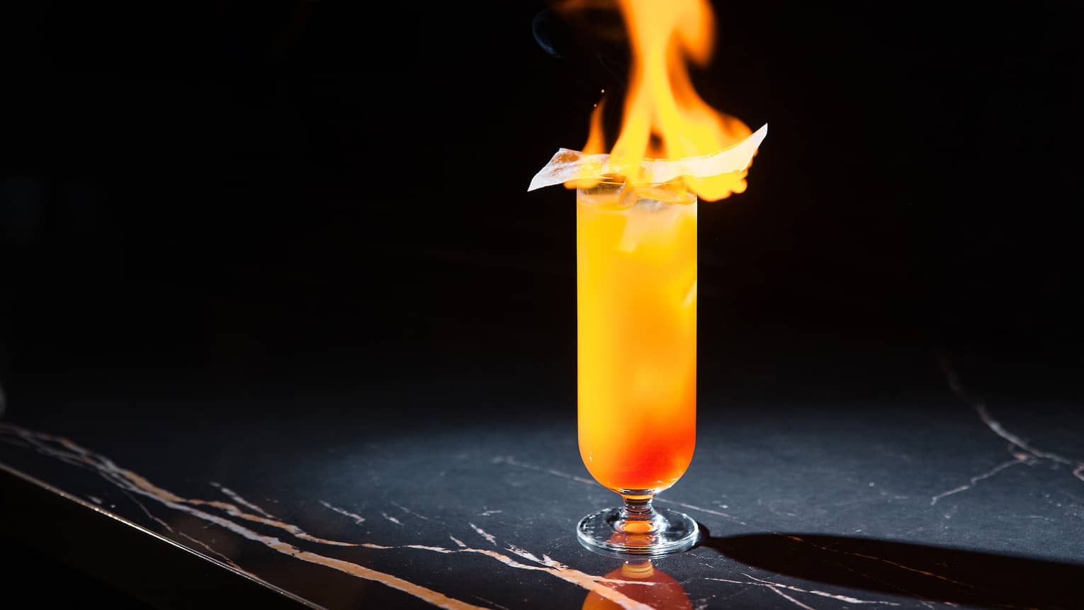 Bright yellow and orange cocktail in tall cylindrical glass, flaming from the top