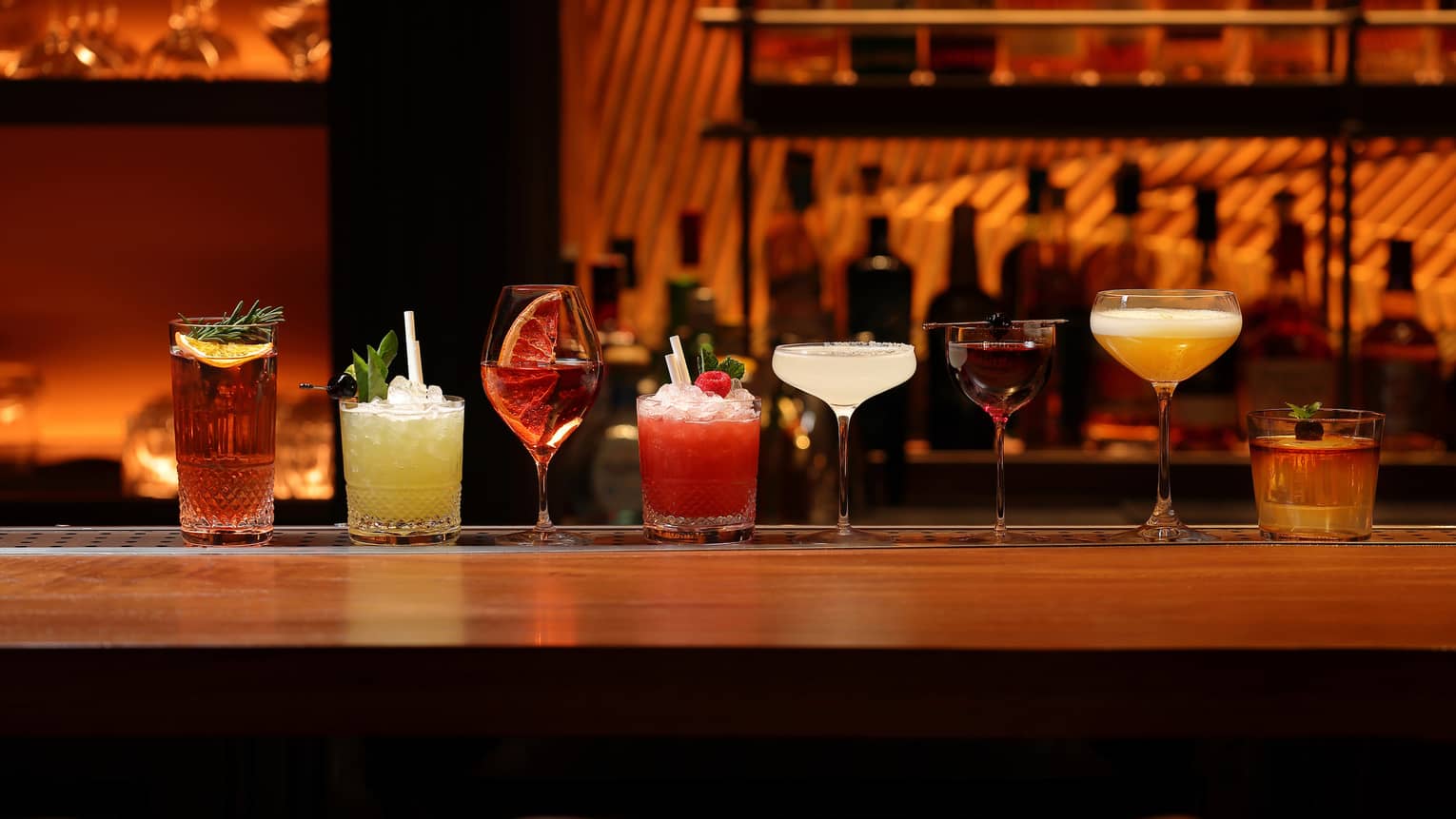 A line of cocktails in various tumblers and stemmed glasses against a backdrop of blurred liquor bottles in reddish lighting.