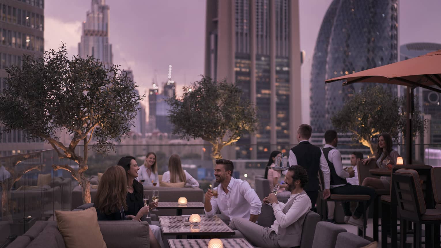 A group of well-dressed people laughing at a metal table overlooking the Burj Khalifa.