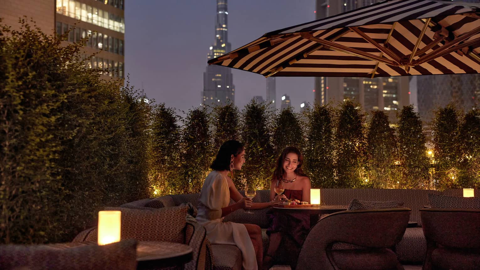 Two women sit at an outdoor table at dusk with a candle in the middle and a patio umbrella overhead