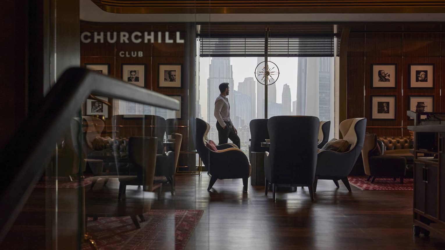Man stands by a floor-to-ceiling window looking out on the city skyline in a dimly lit cigar bar filled with club chairs and leather sofas
