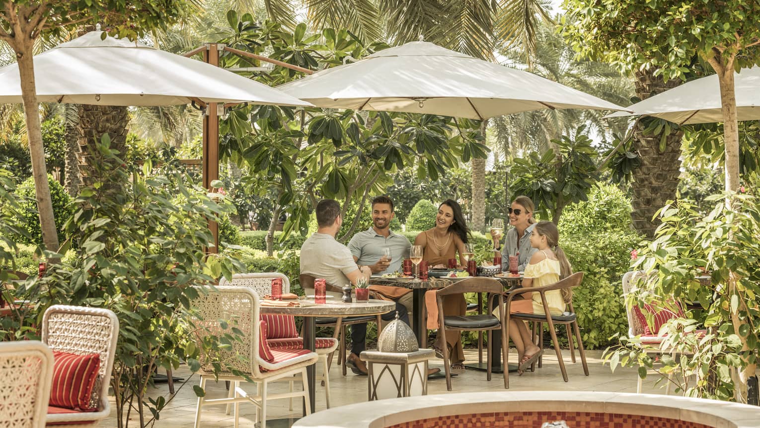 Two couples and a child dine outdoor under an umbrella on SUK's patio surrounded by trees and vegetation