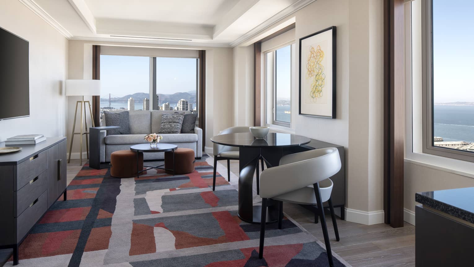 Hotel living room with two grey chairs, carpet with geometric patterns, sofa, TV, windows