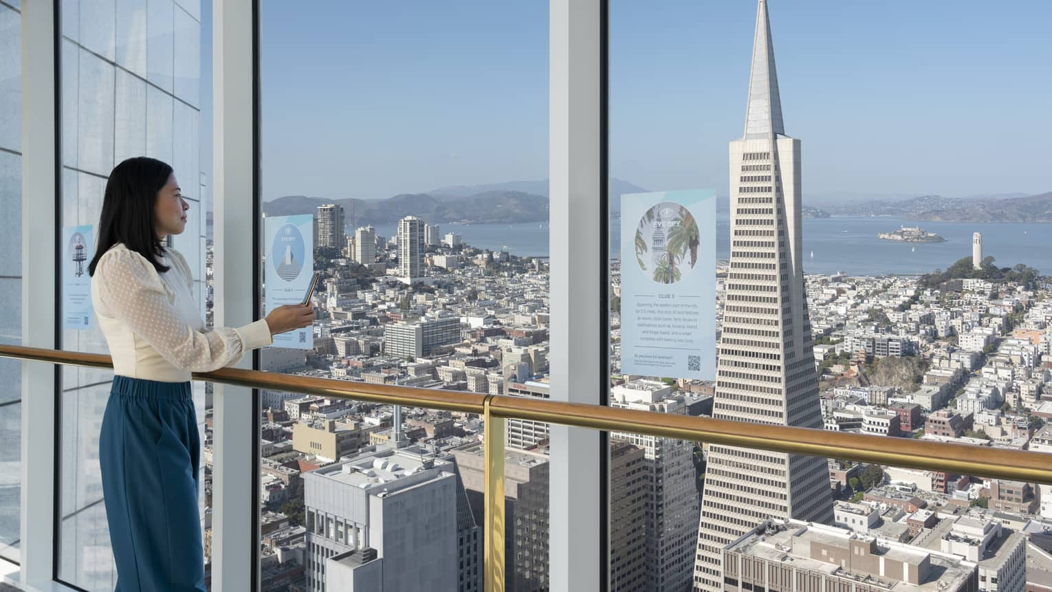 Woman stands inside hotel next to floor-to-ceiling windows, looking out to San Francisco cityscape