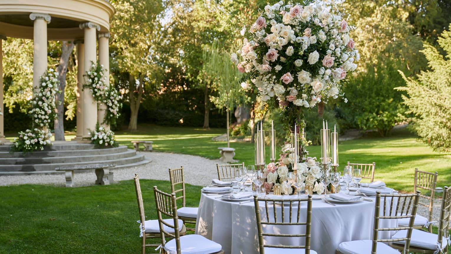 White formally set wedding reception table in garden with large white topiary and gazebo in backdrop