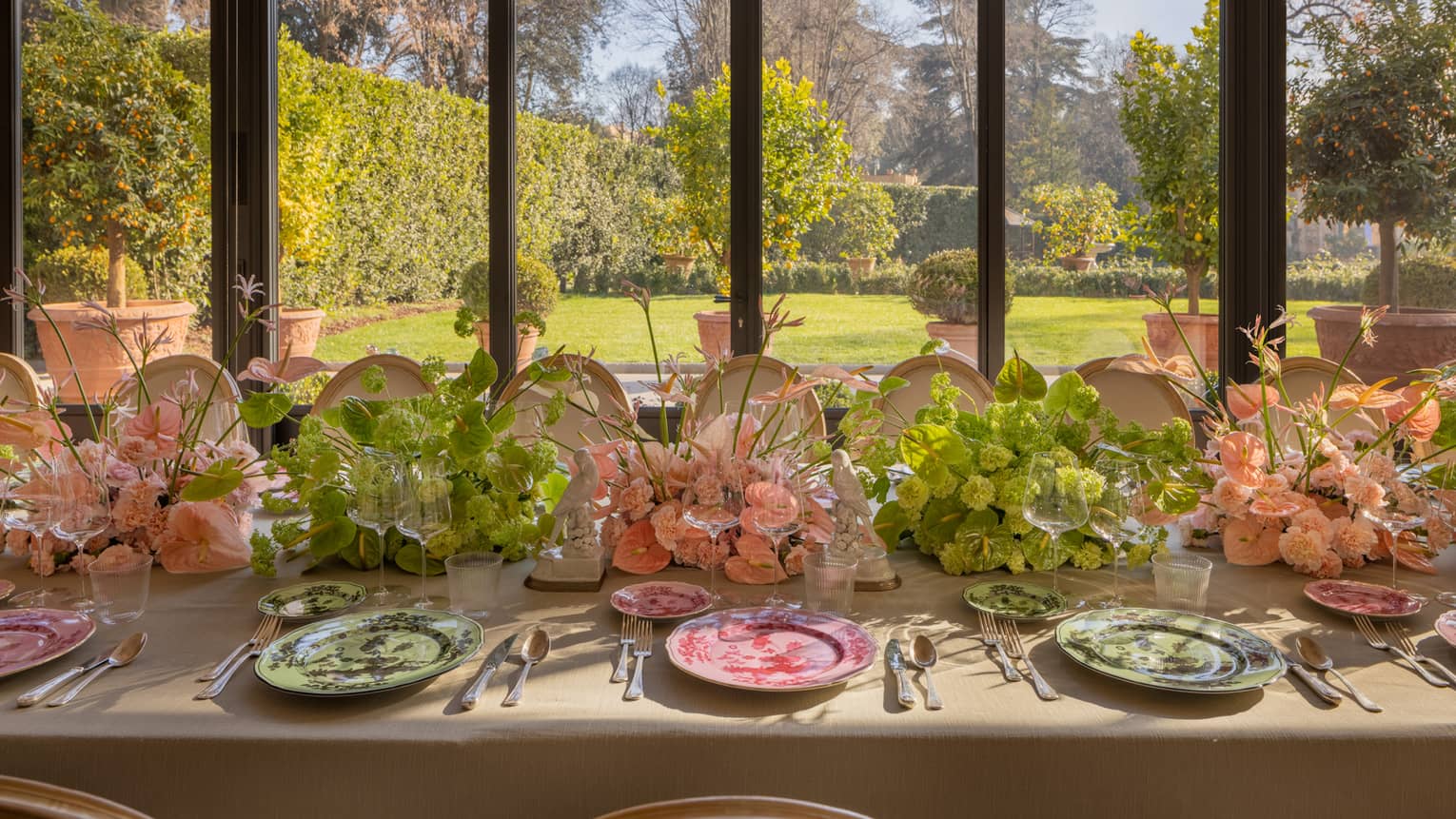 Bright banquet table set with pink and green plate and flowers beside tall windows looking out to manicured garden