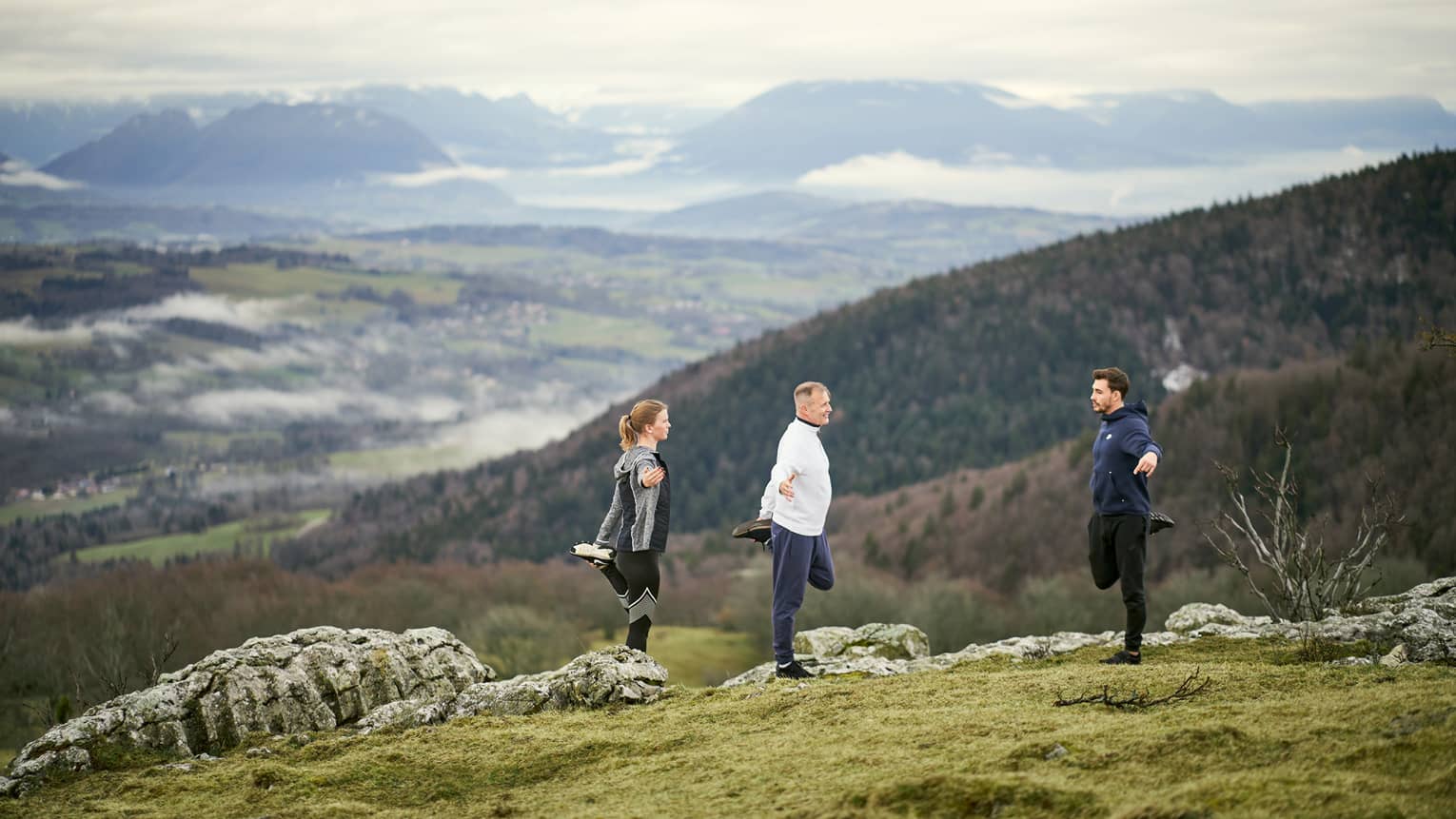 Woman and man standing on mountainside with male trainer, all stretching left quads 
