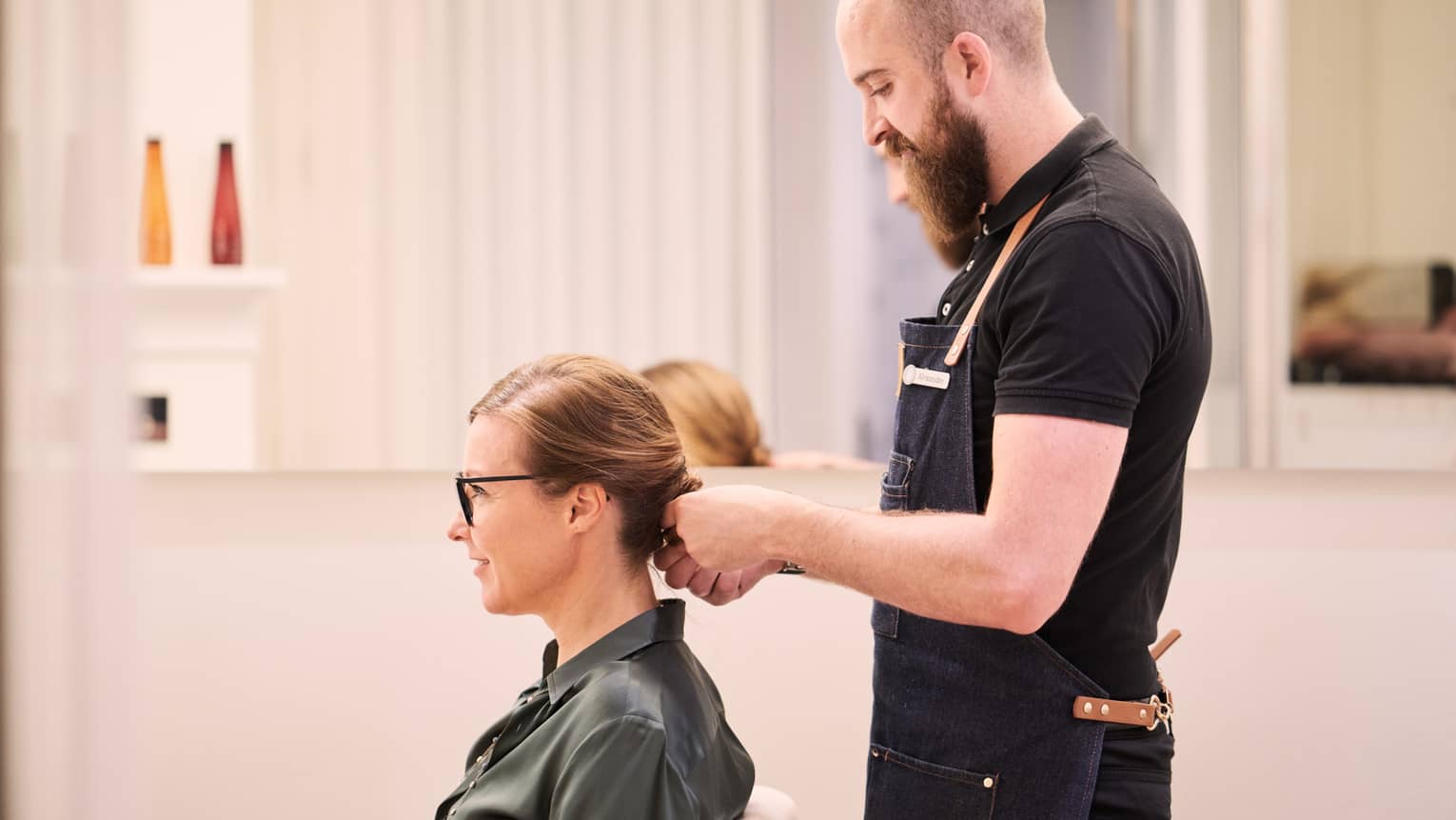 Woman sits in salon chair as hair stylist with long beard holds her hair