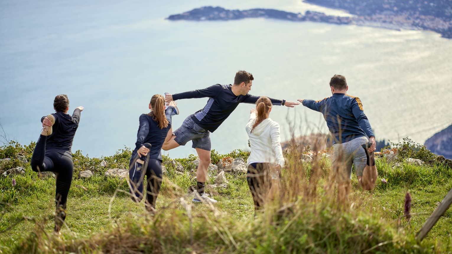 Rear view of four people in yoga poses on grassy cliff with instructor in front, sea view