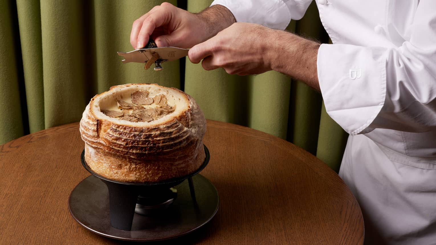 Chef in white chef's coat shaving truffle atop cheese fondue in bread bowl