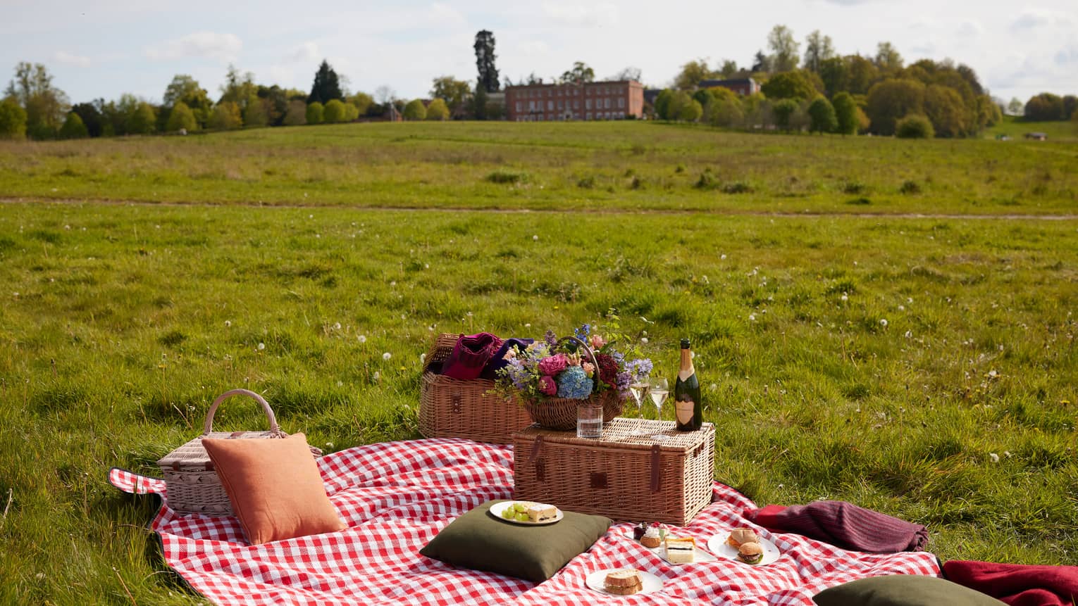 Picnic setup of red gingham blanket with picnic basket topped with flower bouquet and champagne bottle on expansive lawn with large red brick manor in backdrop