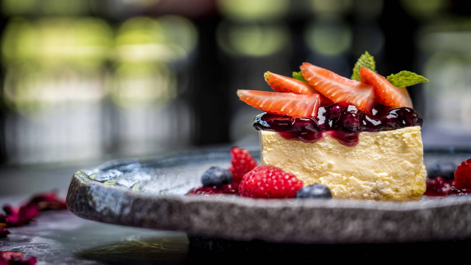 A stone plate of cheesecake topped with berry compote and fresh sliced strawberries, in a bed of fresh berries.