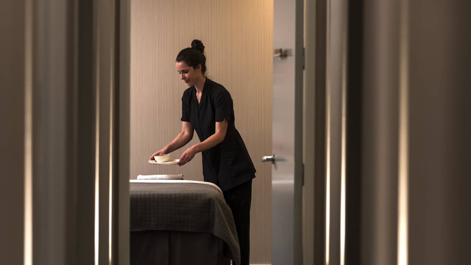 View through door as spa staff sets bowl on massage bed 