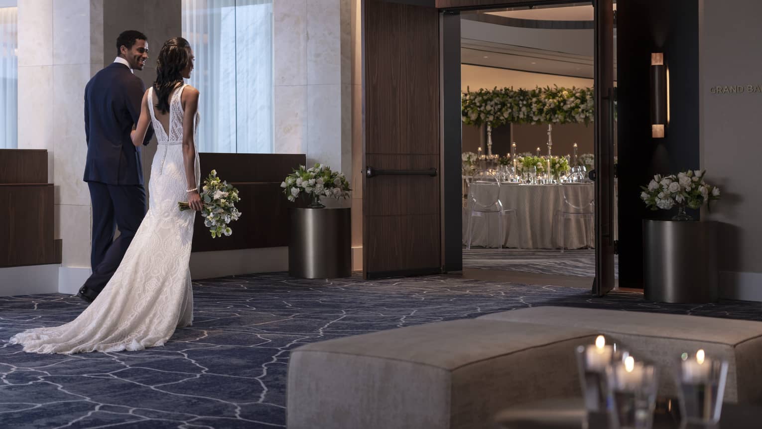 A bridal and groom walking towards the doors of a ballroom, inside the room you can see tables and flowers.
