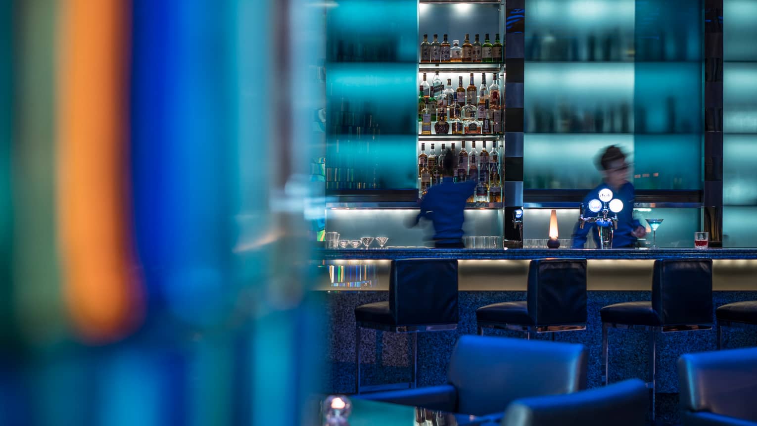 Bartenders behind illuminated blue bar, stools, colourful lights