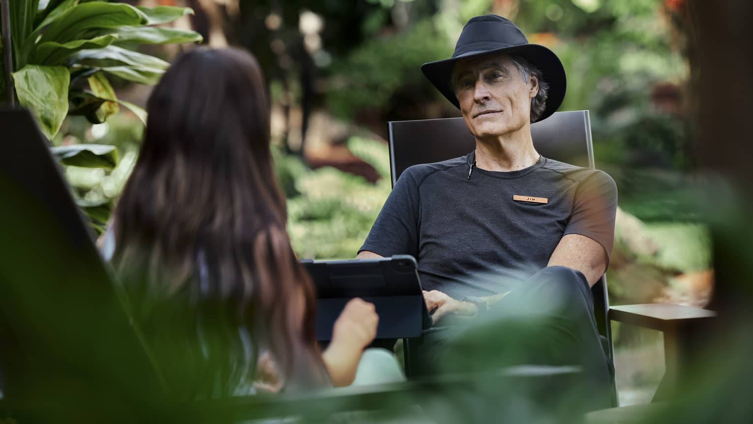 Sitting amid lush vegetation, two people engage in conversation, one with kind eyes and a soft smile typing on a tablet.