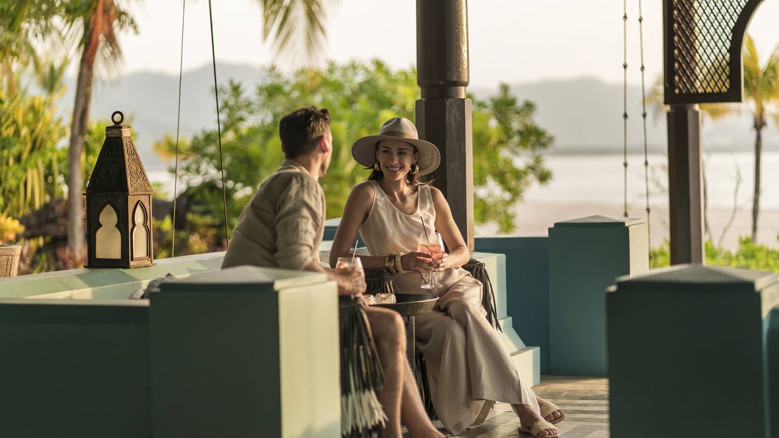 A man and woman are sitting in an outdoor Rhu Bar area overlooking the ocean holding cocktails