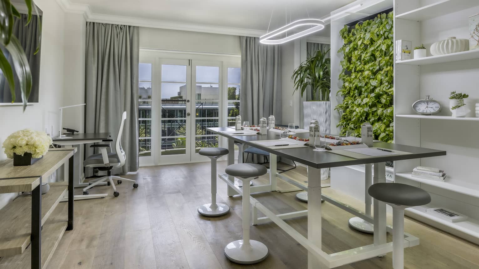 A brightly lit office with a large table, stools, a desk, shelves, and plants on the wall.