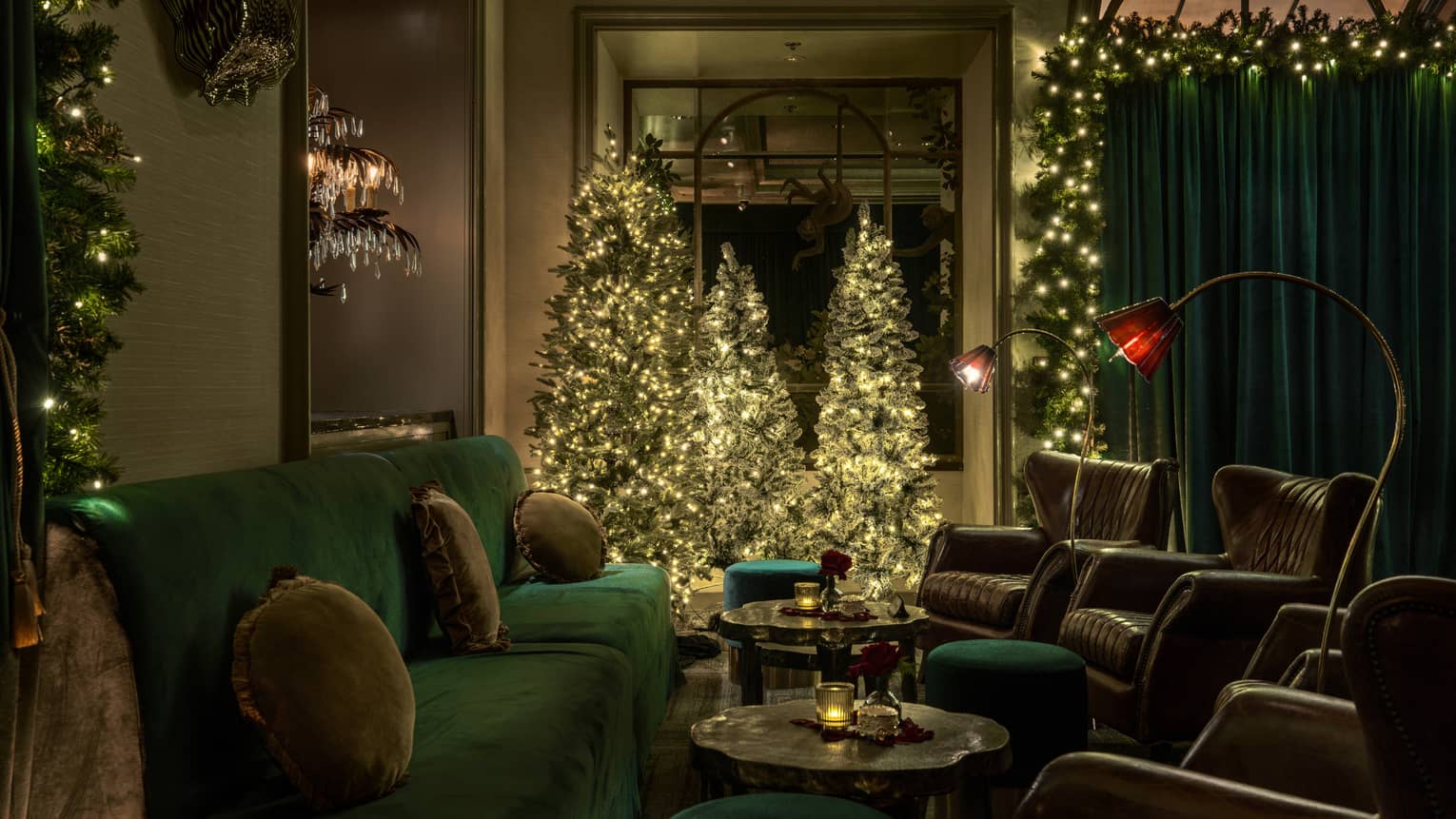 Three Christmas trees sit by a window in a dimly lit lounge filled with green velvet seating and small tables