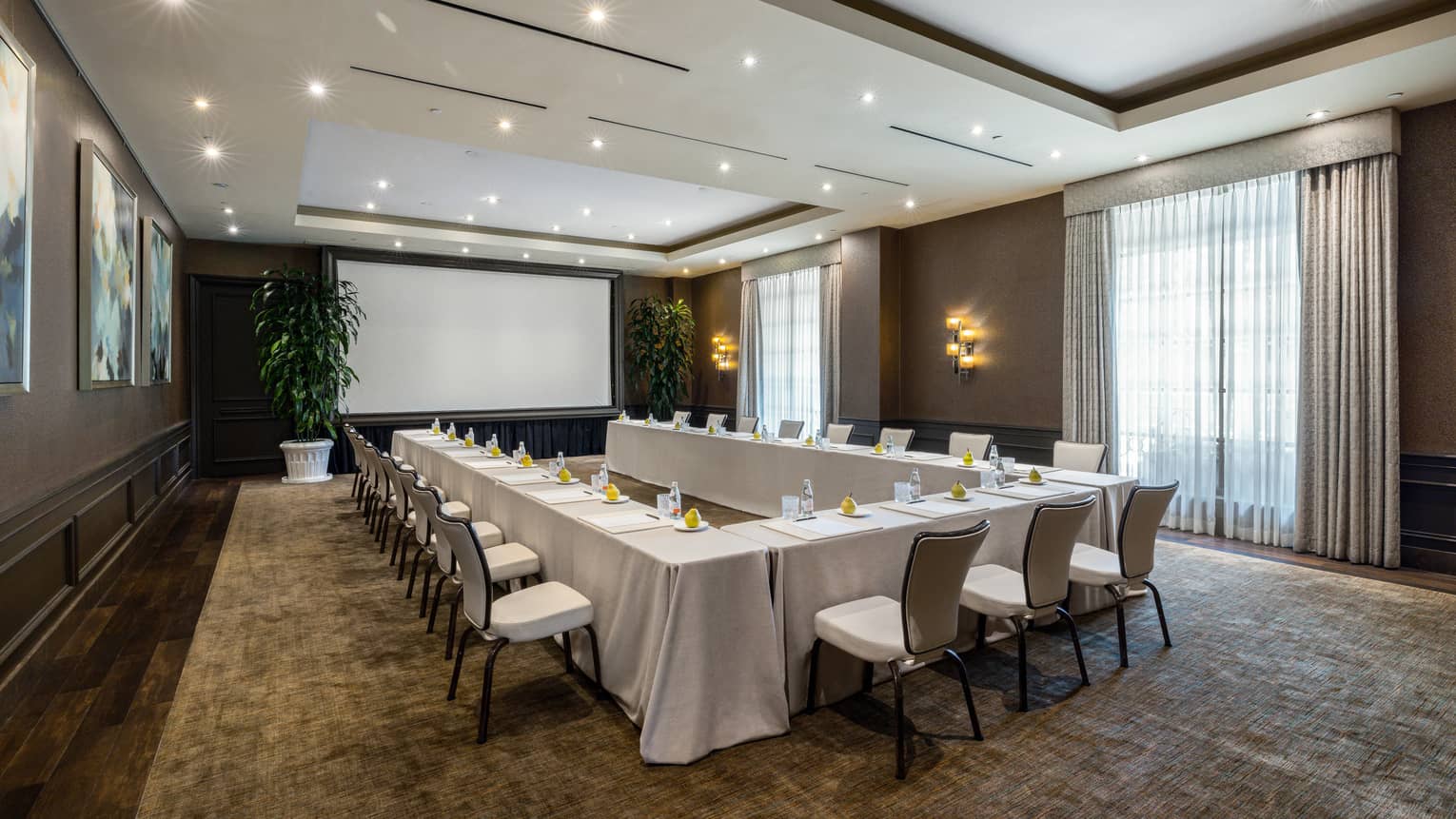 Meeting room set up U-shape style with cream-coloured linens, ambient lighting, projection screen framed by large potted plants, and floor-to-ceiling windows