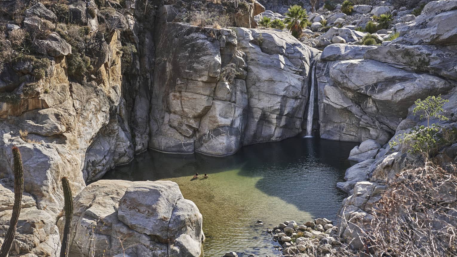Two people wade in a small ocean lagoon surrounded by large cliffs, creating a serene and picturesque scene with clear water and rugged natural beauty.