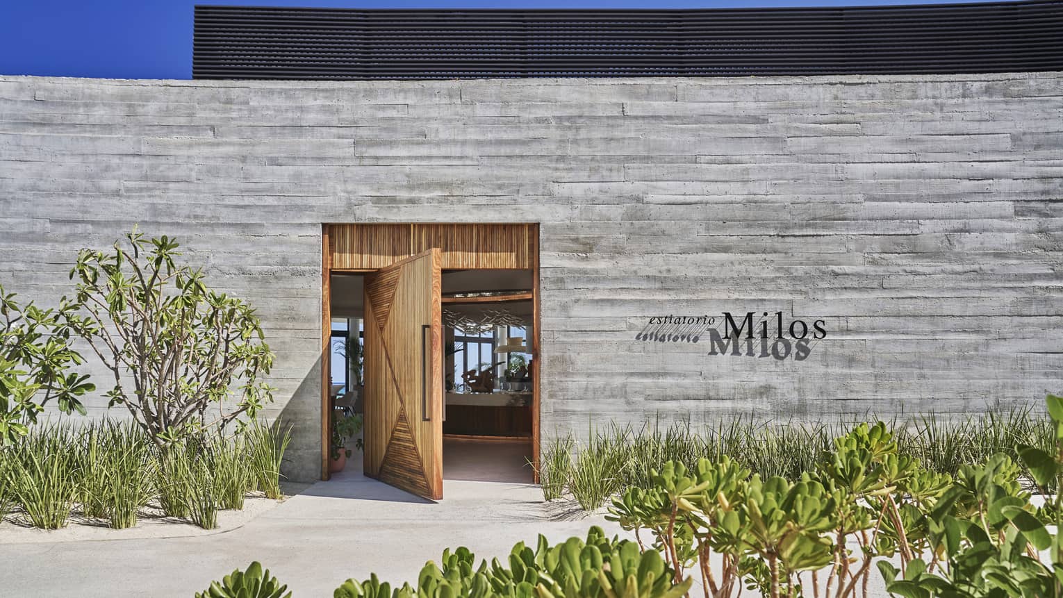 A wood door is open in a grey brick wall, in front of the wall is sand and green plants.