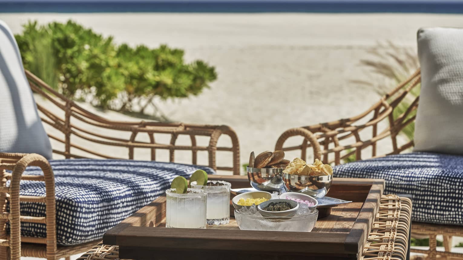 Two cushioned chairs outside with a small table with two drinks, chips and salsa on the beach.