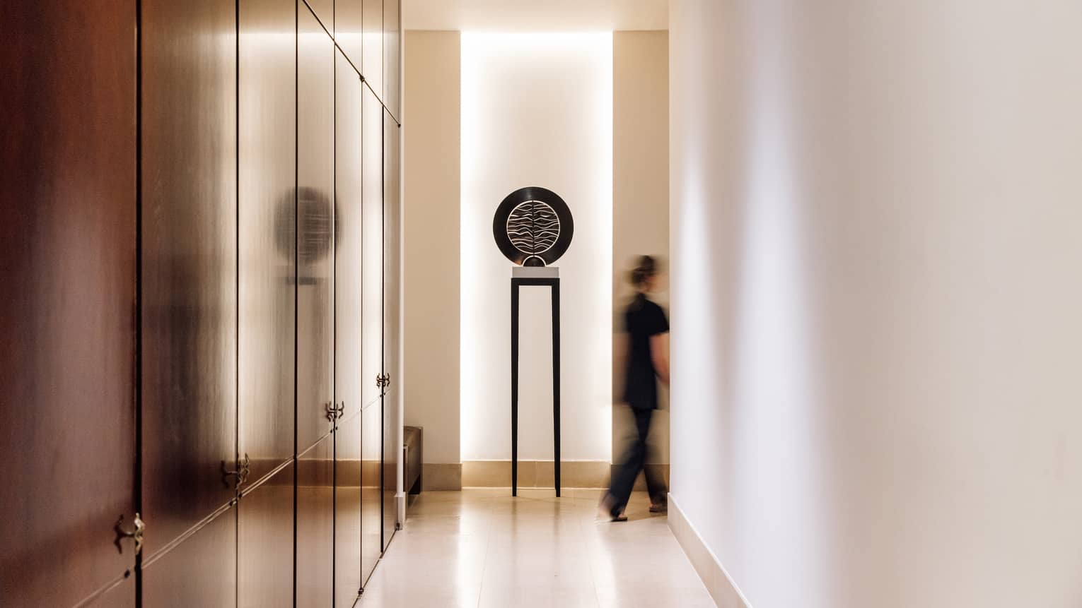 Spa hallway with art piece at the end and a spa therapist dressed in black going through a doorway beside it