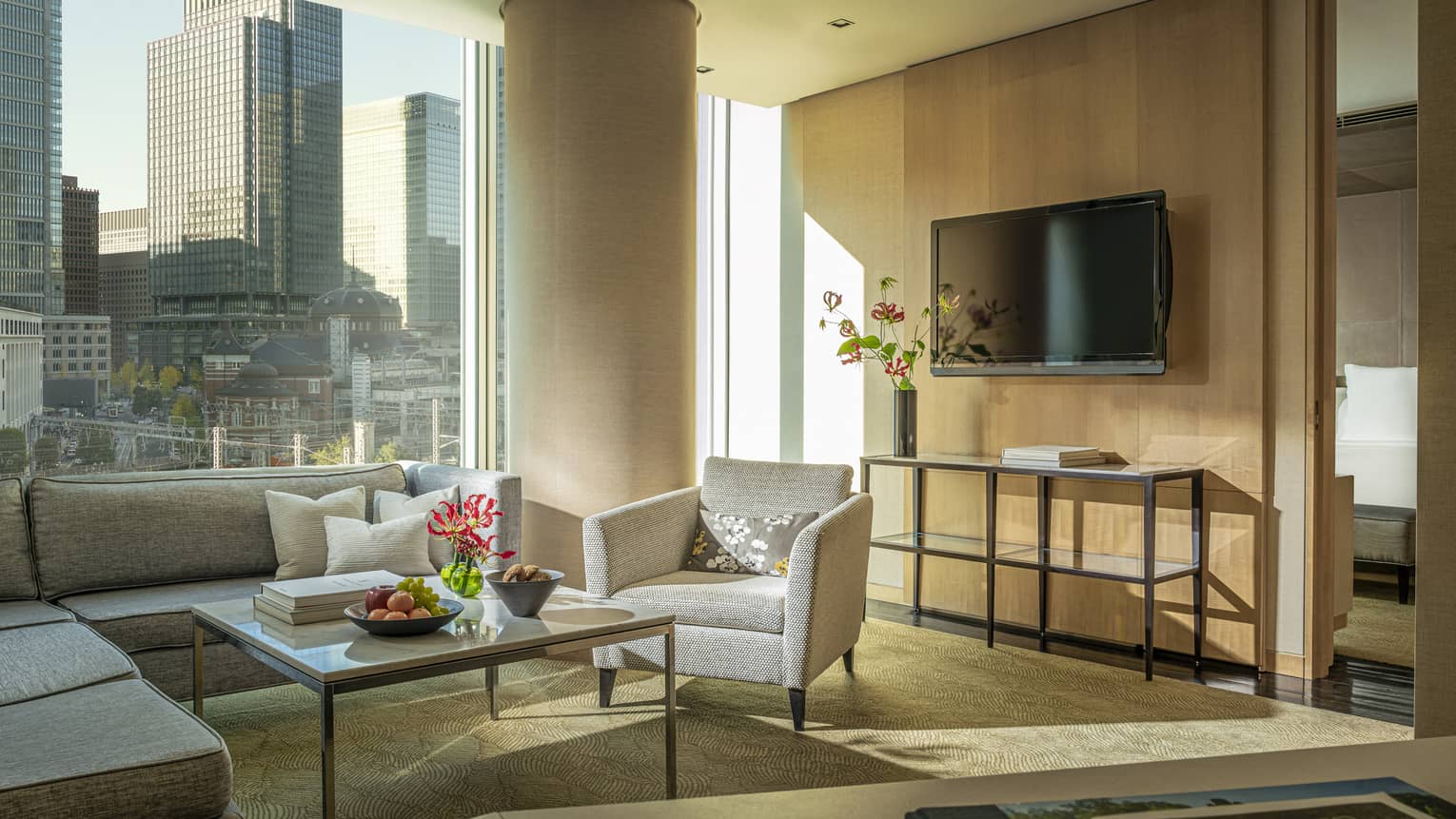Four Seasons executive suite with gray sectional sofa, armchair, coffee table with fruit and flowers, large window with city view, TV and console table 