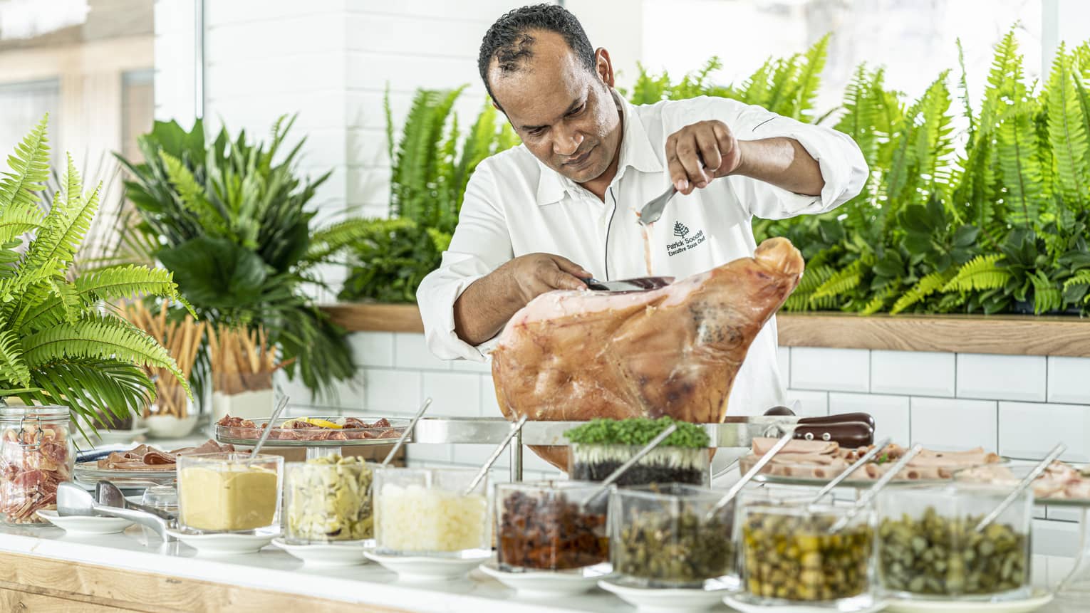 Chef in white shirt slicing meat on counter behind row of condiments in dishes