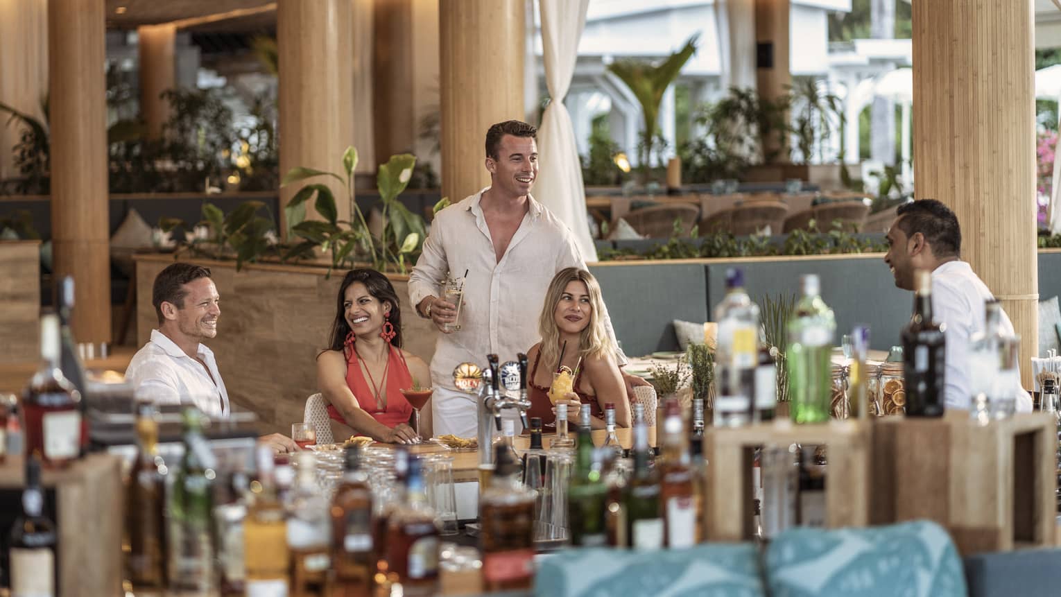 Four friends smiling while having drinks and talking to bartender behind stocked bar