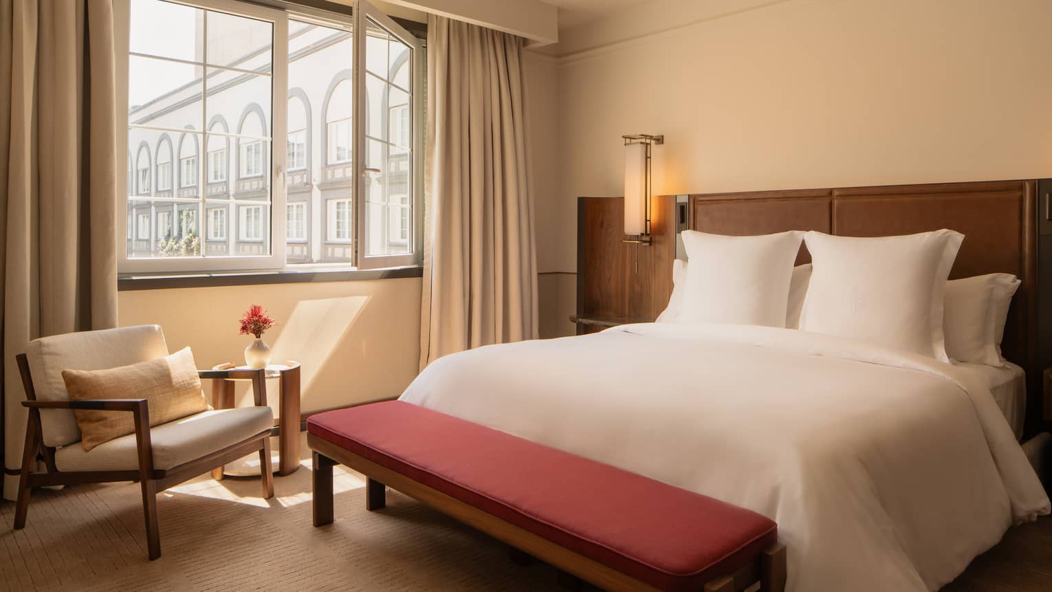 Hotel room with king bed, red bench, arm chair and window looking out to courtyard