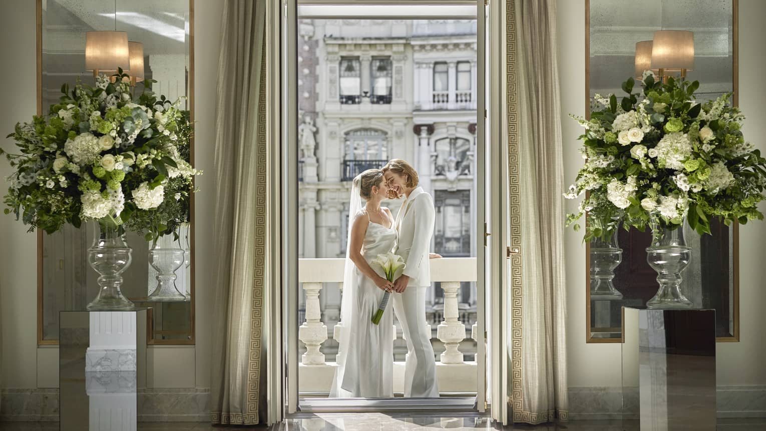 Wedding couple dressed in white standing intimately on terrace, framed by doorway with tall interior curtains, framed by large white flower arrangments