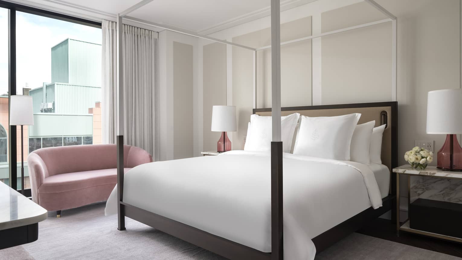 A modern hotel bedroom houses a four-poster bed, white linens and pink accent lamps. A pink loveseat sits near large windows, offering views of adjacent buildings.