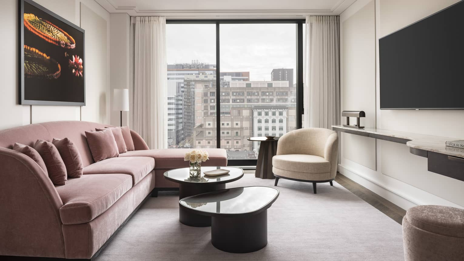 A seating area featuring a pink L-shaped couch, a circular white loveseat, and an cityscape view from a full-length window.