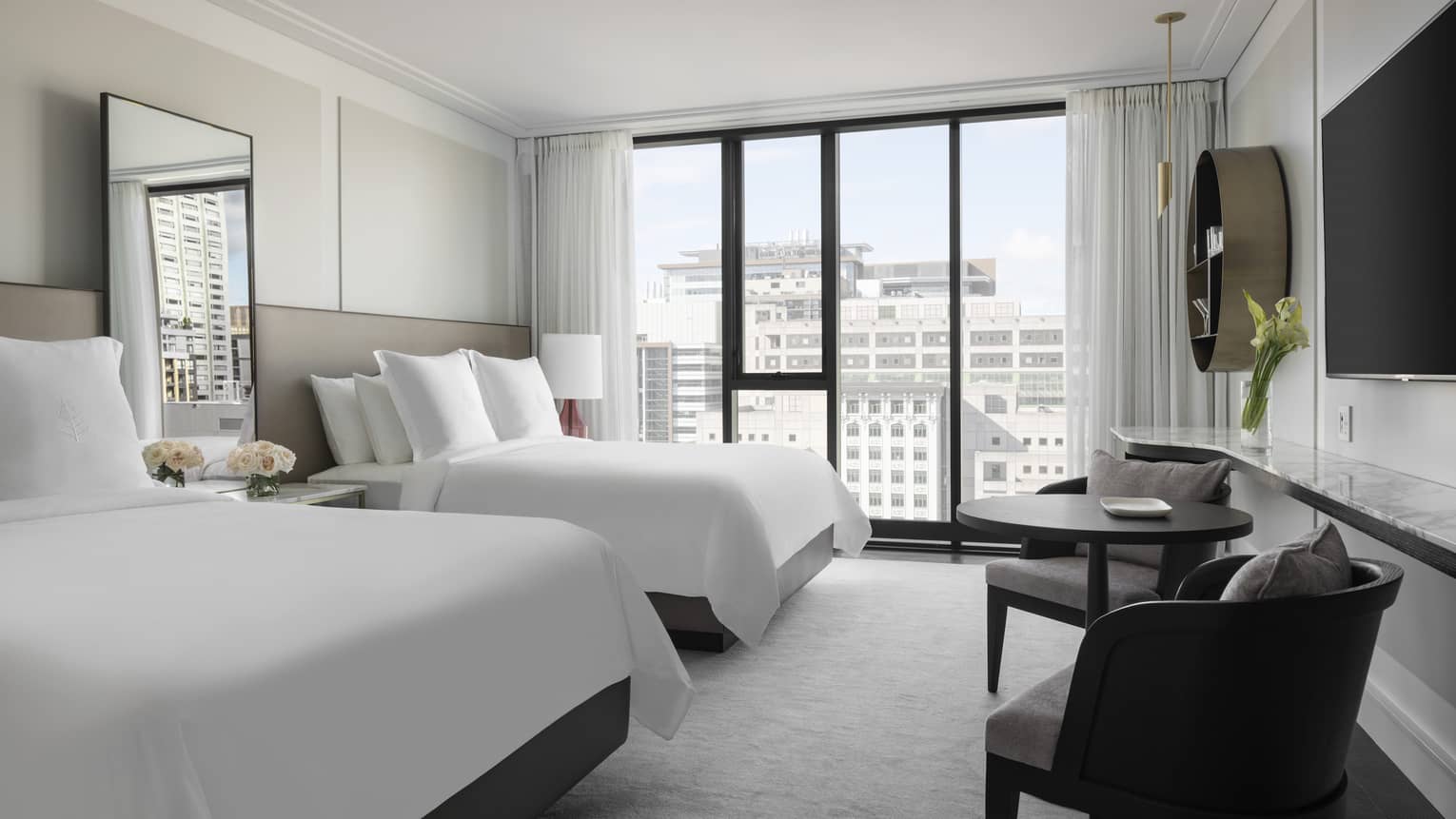 A bright hotel room with two double beds featuring white linens, large windows with city views, wall-mounted TV, and small seating area with table and chairs