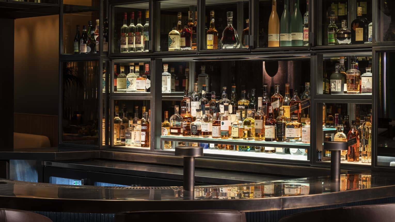 Dark leather seats frame a stainless-steel counter in front of lit-up mirrored shelves fully stocked with premium liquor. 