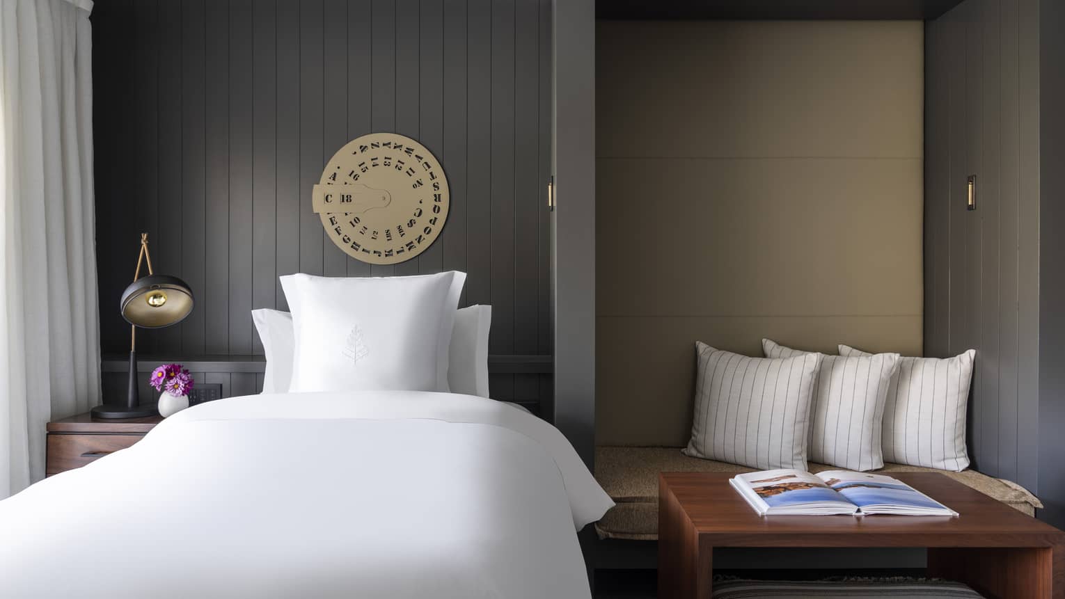 Luxury resort room with white bed, dark paneled walls, decorative wall art, bedside table with lamp and flowers and seating nook with striped pillows and book