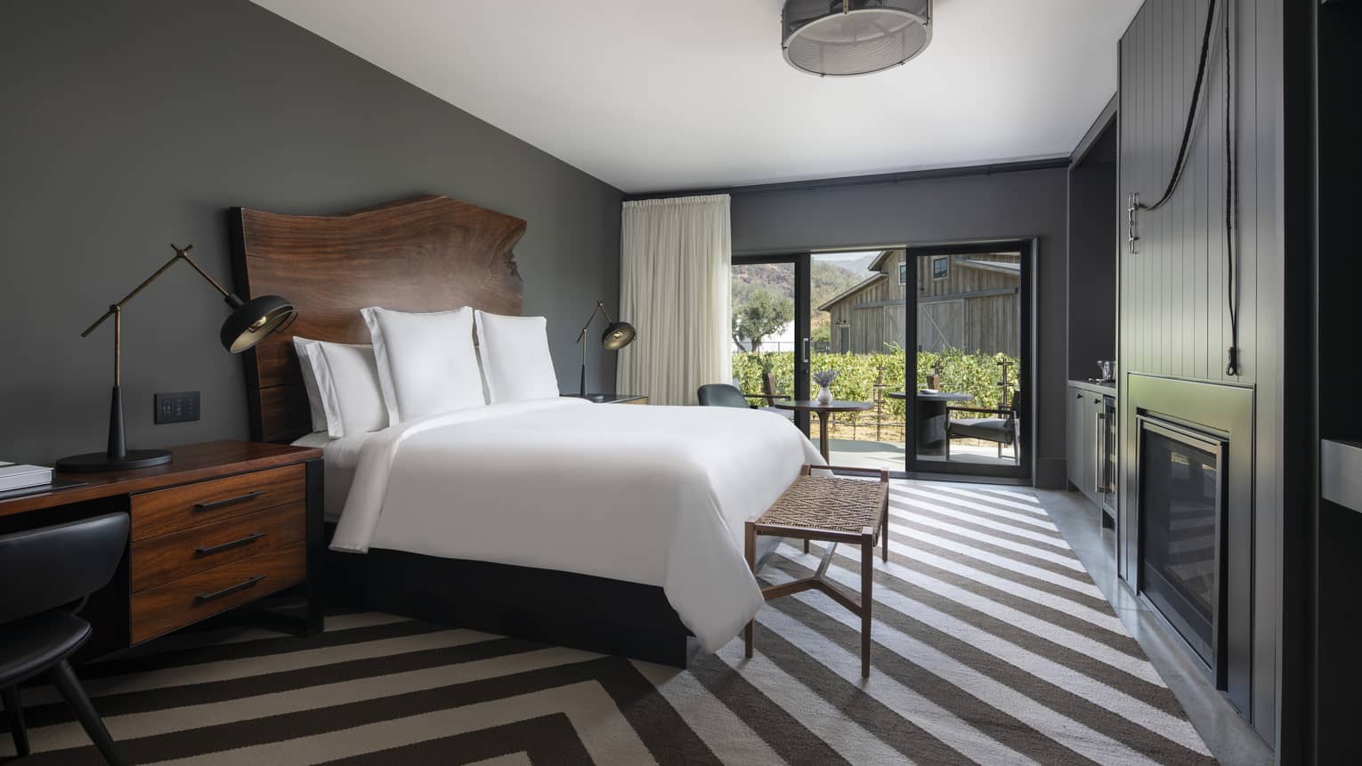 Luxury resort room with a king bed, wooden headboard, bedside lamps, desk, striped rug, fireplace and patio with vineyard and rustic barn view
