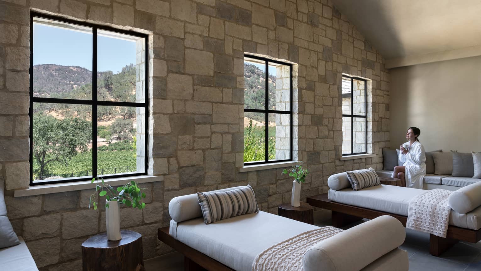 Woman in spa robe sits on couch with a tea in hand in large sunny treatment room at Spa Talisa, looking out window at Napa Valley hillside
