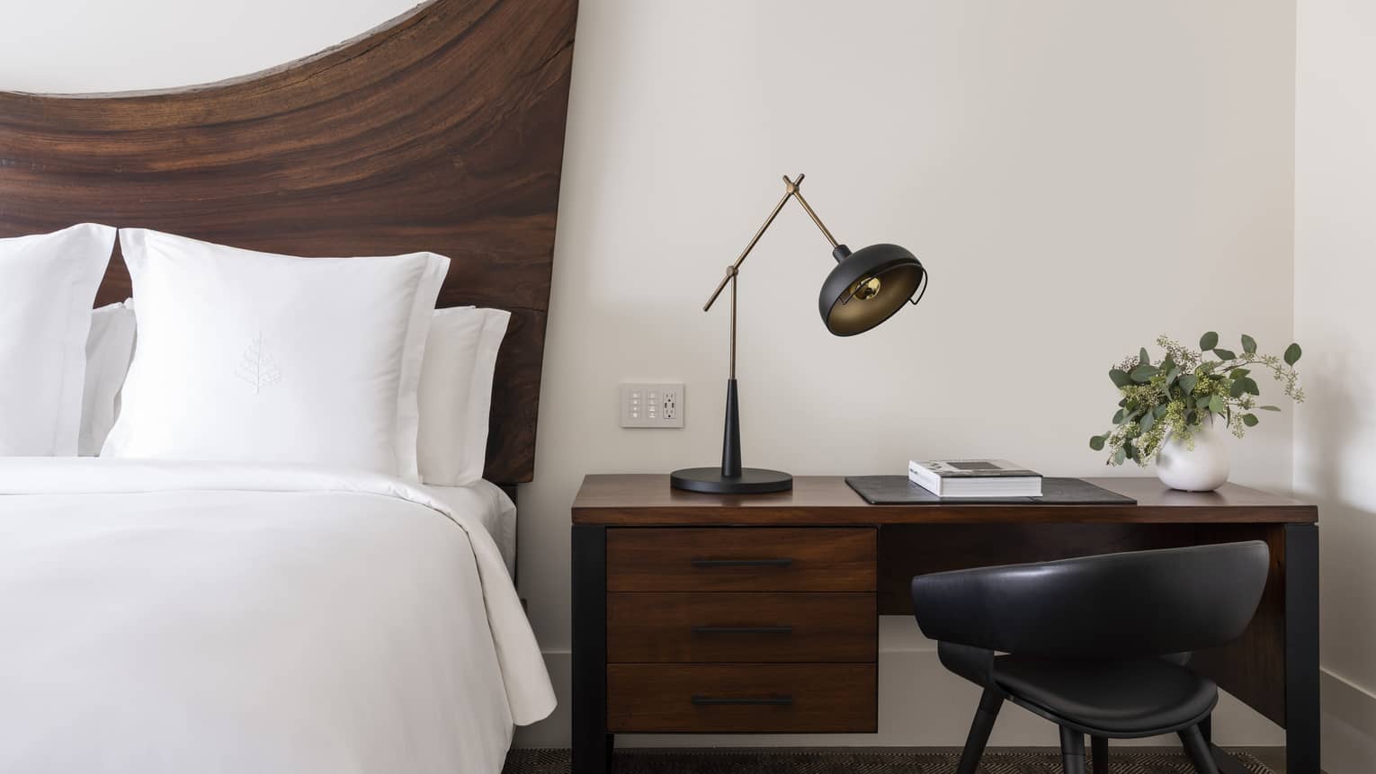 Close-up of a luxury resort king bed with wooden headboard, dark wood desk, black lamp, black chair, book and white vase with greenery