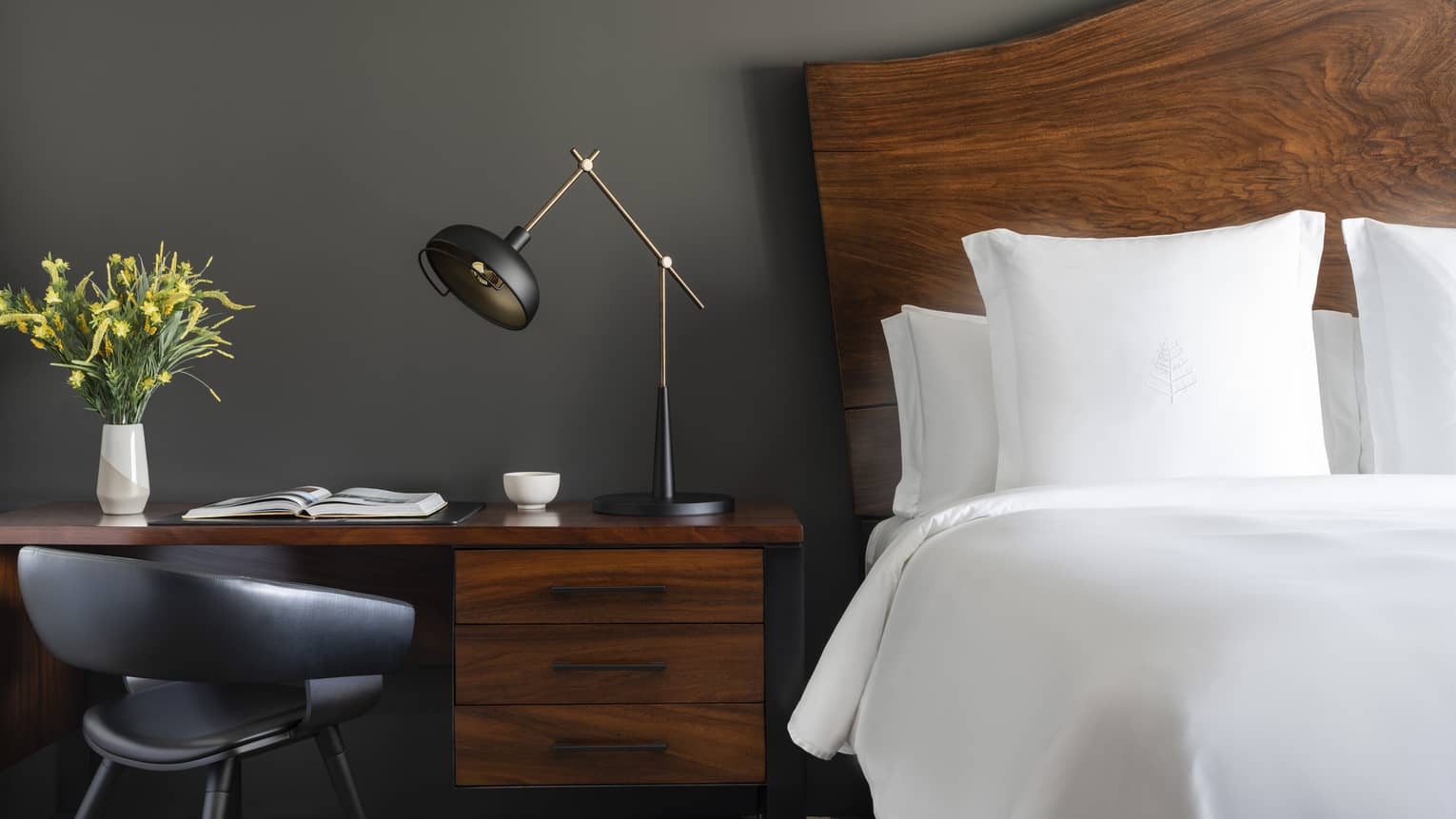 Close-up of a luxury resort white king bed with a dark wood headboard, wooden desk with an open book, modern black lamp, gray chair and vase of yellow flowers