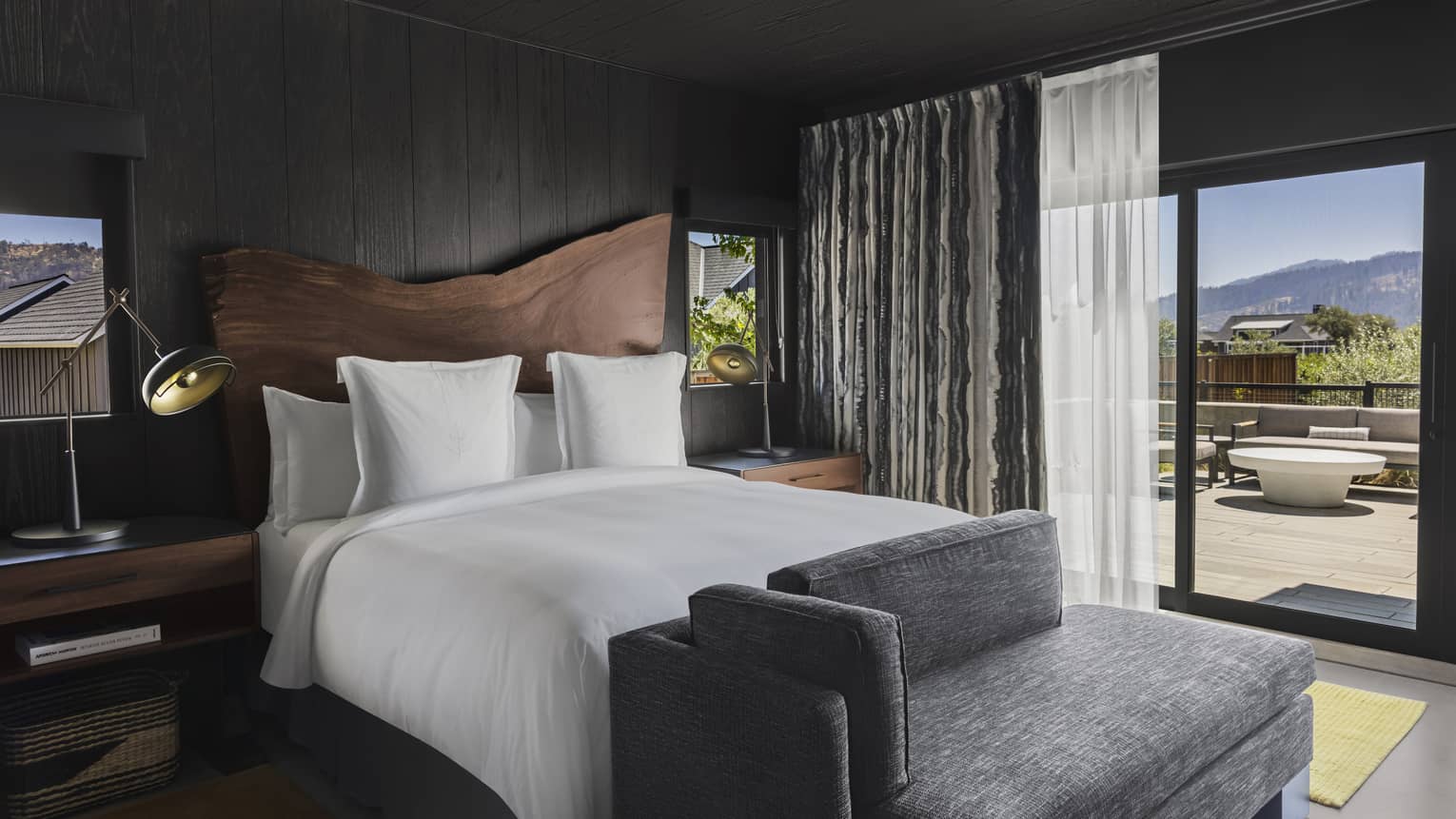 Bedroom with dark wood paneling, king bed, wood headboard, gray chaise, nightstands with lamps and glass doors leading to a private mountain-view terrace 