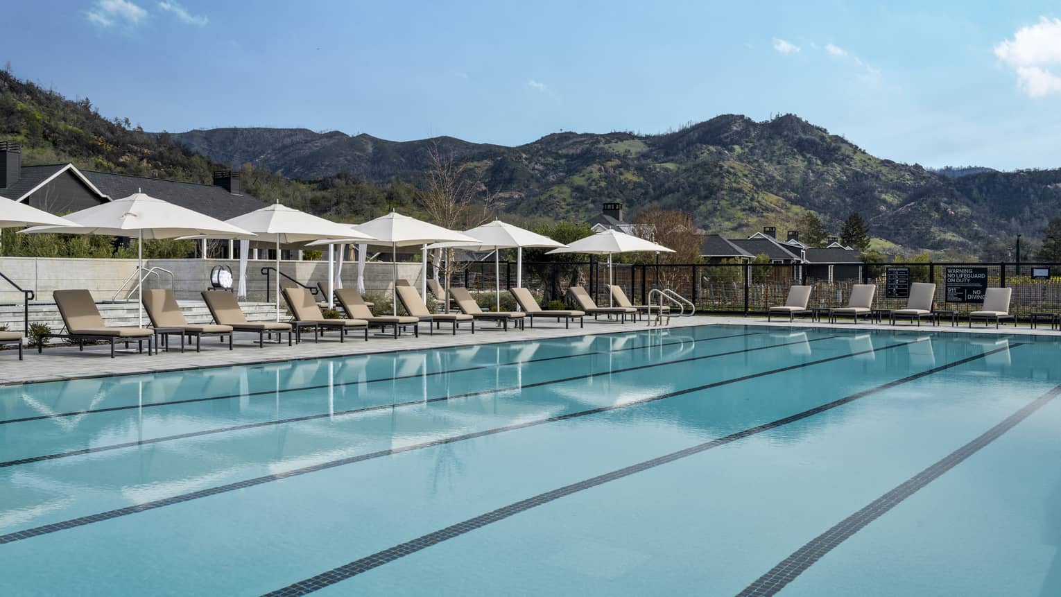 Lounge chairs and umbrellas line the deck around an empty pool surrounded by grassy peaks stretching toward a clear blue sky.