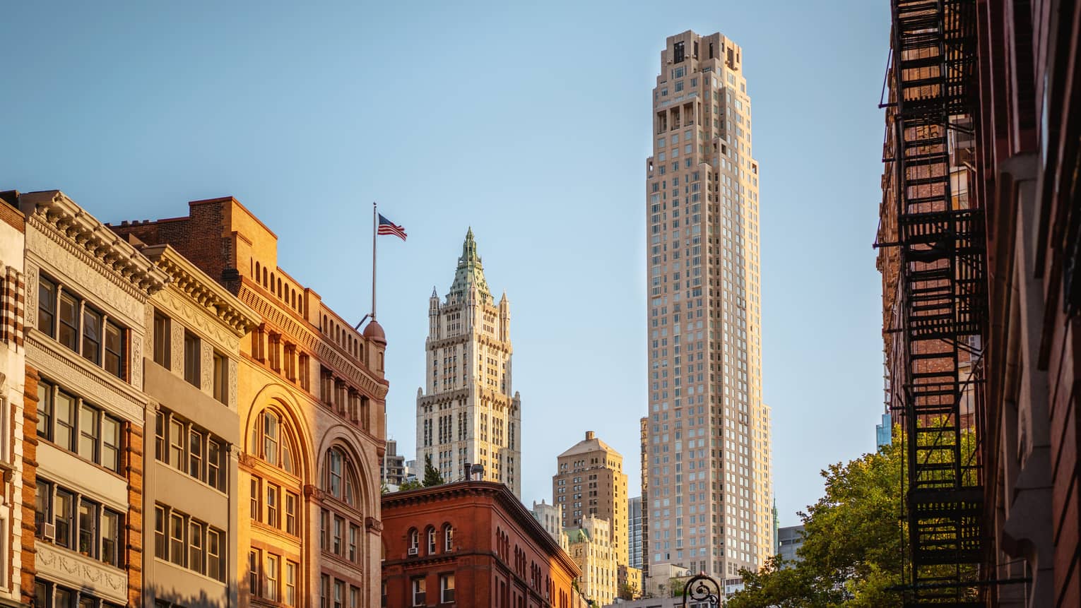 Downtown cityscape view featuring historic and modern skyscrapers, including the Woolworth Building, with an American flag against a clear blue sky