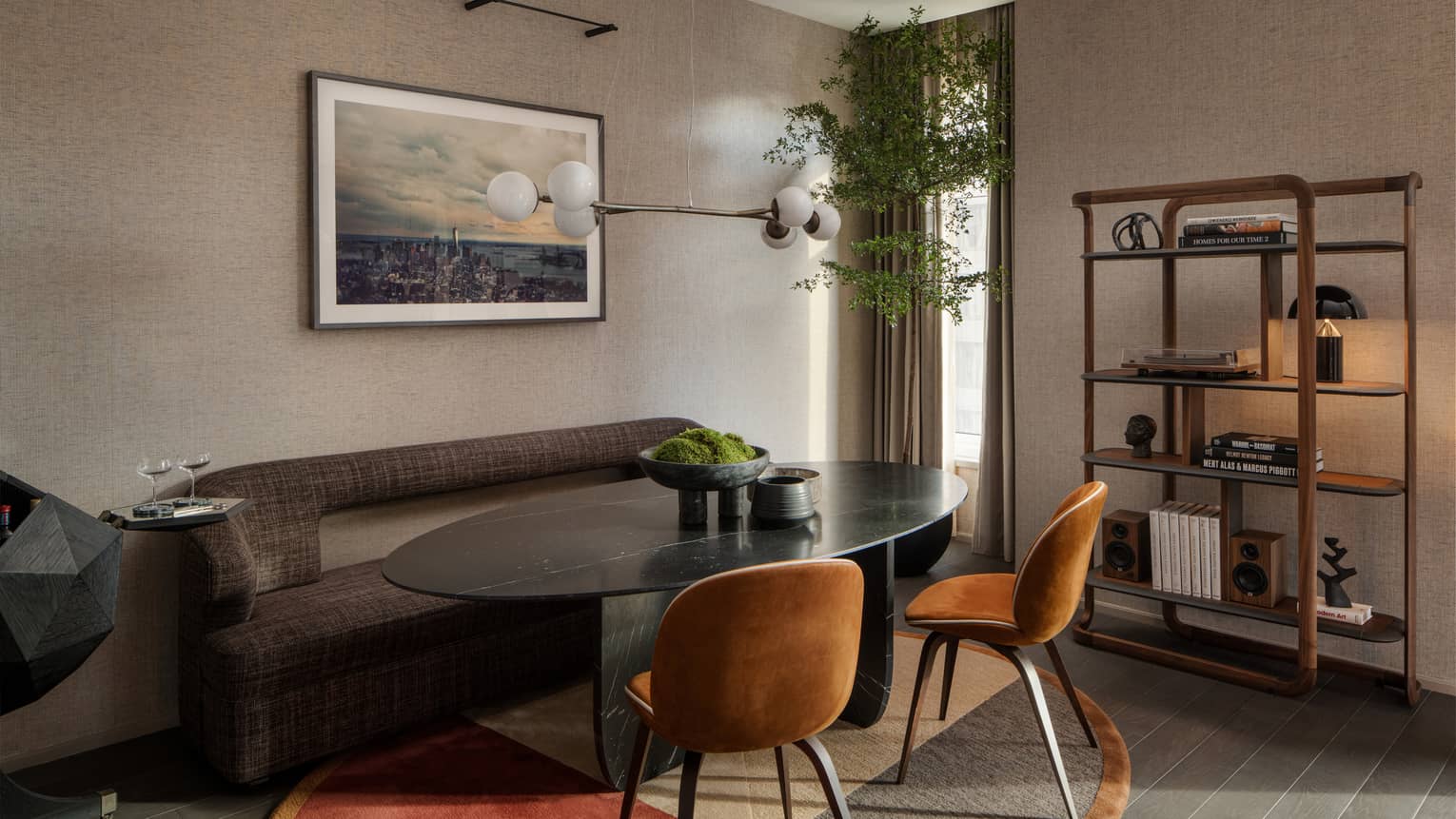 Luxury suite dining area with a dark oval table, brown chairs, gray sofa, cityscape photograph on the wall, modern chandelier and bookshelf with décor items